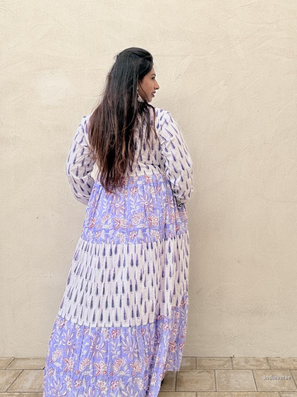 Showing the backside of the dress a woman standing in front of a beige wall wearing a beautiful long cotton dress with floral and cypress prints in aquamarine blue tones