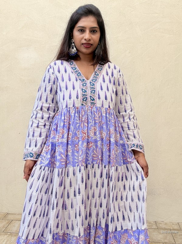 Woman standing in front of a beige wall wearing a beautiful long cotton dress with floral and cypress prints in aquamarine blue tones