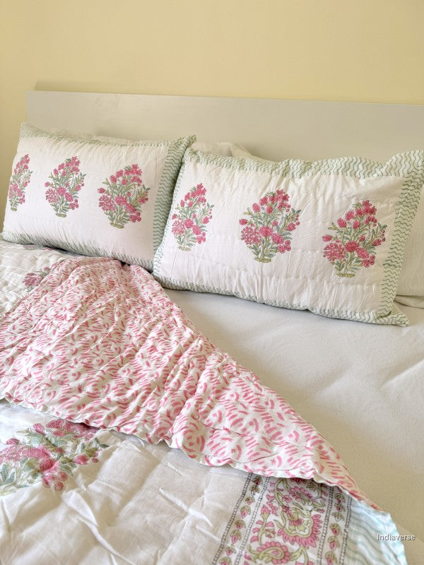 Two pillowcases with pink floral design arranged on a bed with white a bed sheet and a semi-folded quilt with the same floral pattern on a bed