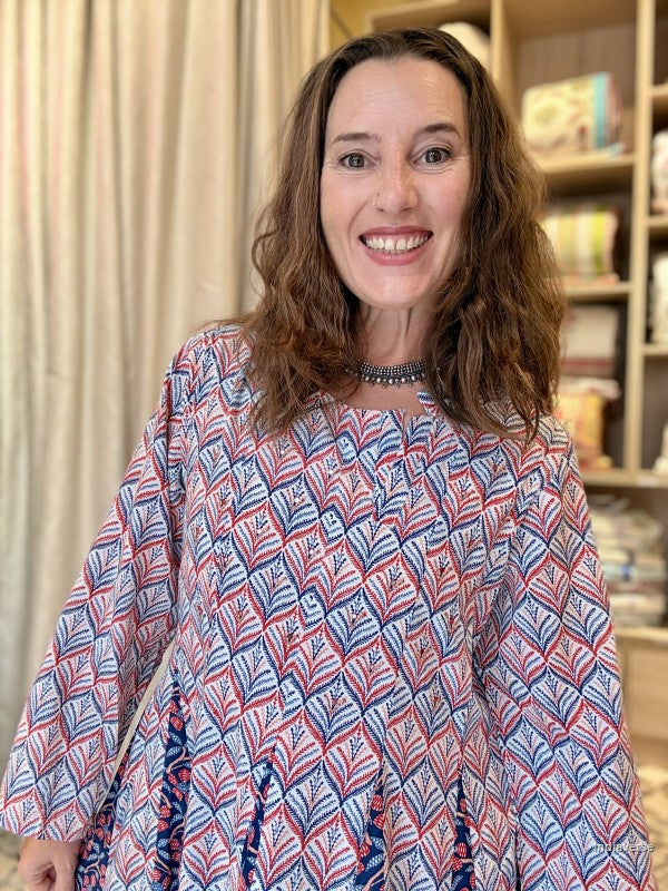 A smiling woman with wavy brown hair wearing a long-sleeve tunic top with a red, white, and blue geometric pattern, standing in front of a bookshelf and curtains. | blue