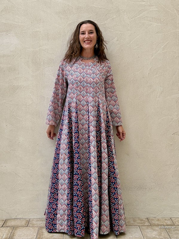 A woman wearing a long-sleeved maxi dress with a pink, blue, and white geometric pattern, standing against a textured beige stucco wall | blue