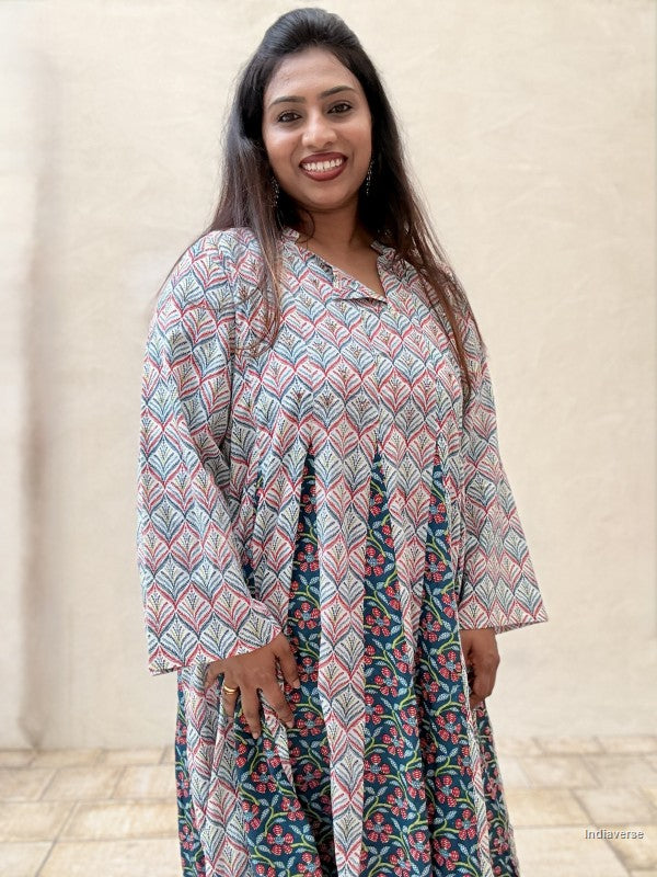 A woman wearing a long-sleeved, V-neck dress with a pink, teal, and white geometric pattern on the upper half and a dark teal floral print on the lower half, standing against a beige stucco wall.