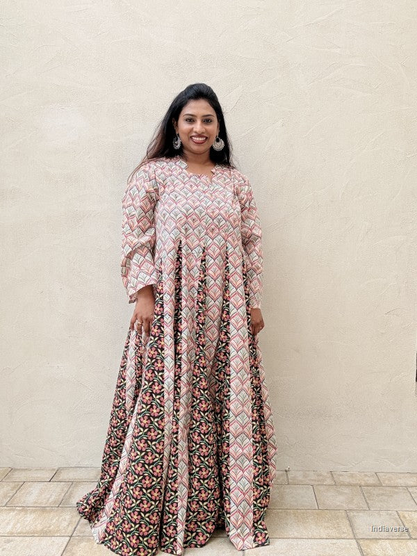 A woman modeling a long-sleeved maxi dress with a geometric pink, beige, and black pattern on the bodice and a floral print on the skirt, standing against a textured beige wall.