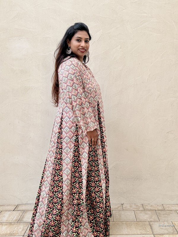 A woman wearing a long-sleeved, floor-length printed dress with geometric and floral patterns in pink, black, and beige, standing against a textured beige stucco wall.