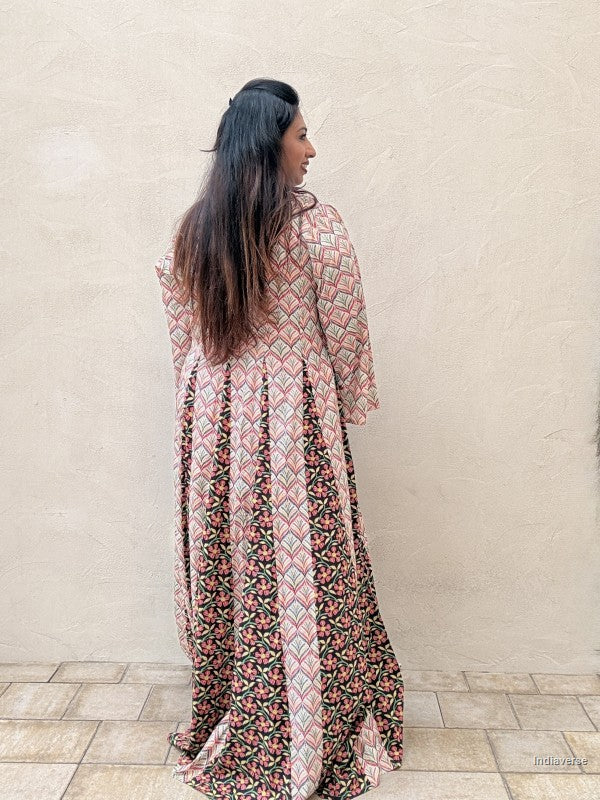 A woman with long dark hair is shown from behind wearing a long-sleeved maxi dress with a geometric and floral print. The dress features a dusty pink base with green, black, and white patterns, set against a neutral beige stucco wall and tiled floor.