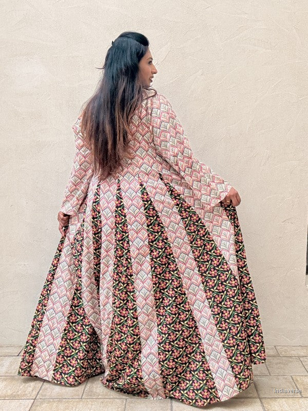 A woman wearing a long-sleeved, floor-length printed dress, photographed from behind as she holds out the sides of the skirt to show its volume and pattern. The dress features vertical panels of pink and beige geometric patterns and dark green floral prints.