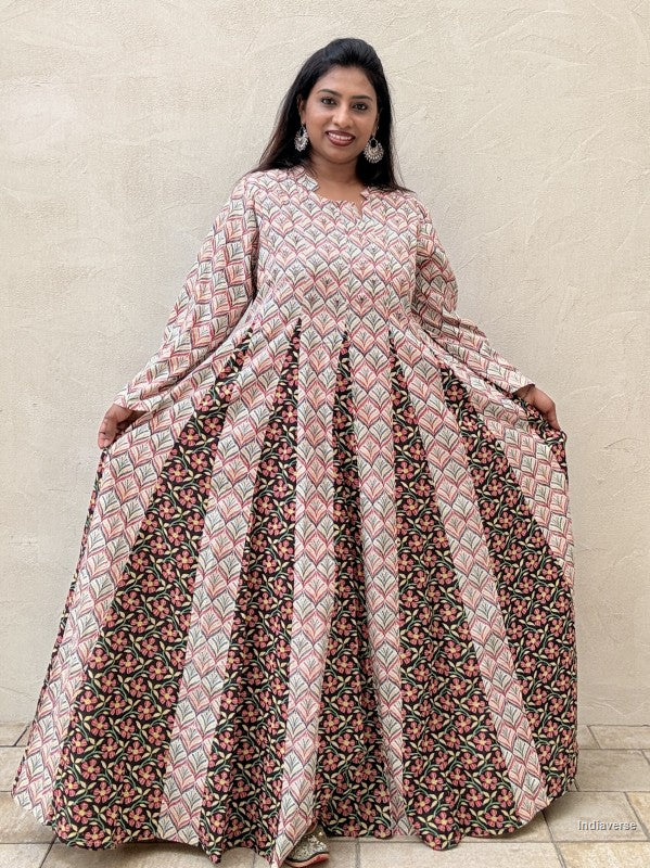 A woman modeling a long-sleeved, floor-length Anarkali gown with a geometric pink, beige, and black pattern on the bodice and pleated skirt, set against a neutral beige wall.