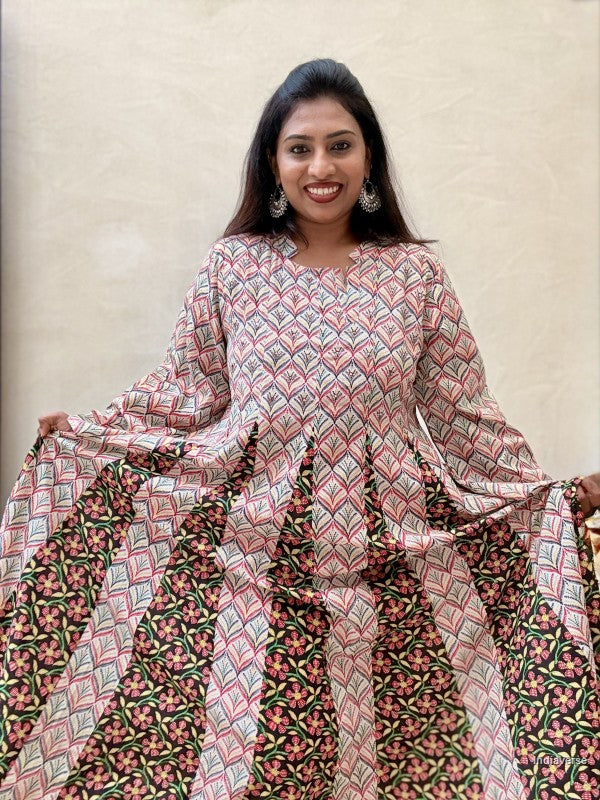 A woman modeling a long-sleeved, wide-leg kurta with a geometric pink, green, and black block print pattern, holding out the skirt to show its full drape against a neutral beige background.