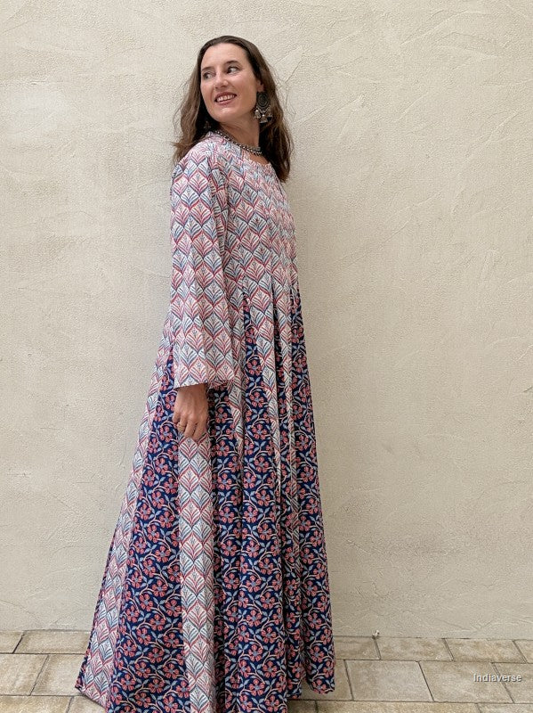 A woman wearing a long-sleeved maxi dress with a pink, blue, and white geometric and floral pattern, standing against a textured beige stucco wall. | blue
