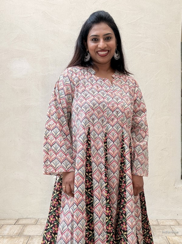 A woman wearing a long-sleeved printed kurta dress with a geometric pattern in pink, beige, and black, standing against a textured beige wall.