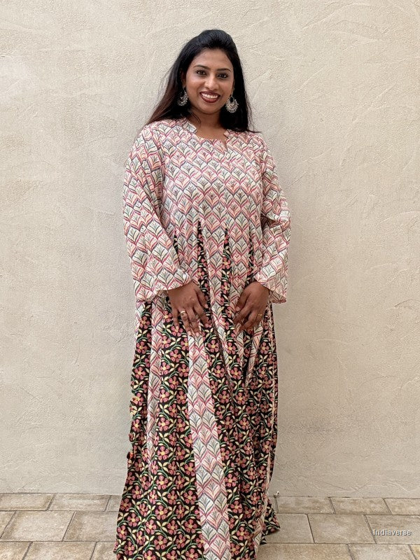 A woman modeling a long-sleeved maxi dress with a geometric and floral block print pattern in pink, black, and green, standing against a textured beige wall.