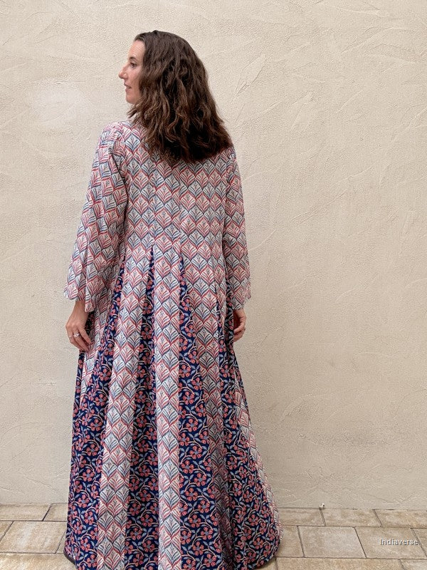 A woman with long brown hair is shown from the back wearing a long-sleeved maxi dress with a geometric and floral block print in pink, blue, and white. She is standing against a textured beige stucco wall.