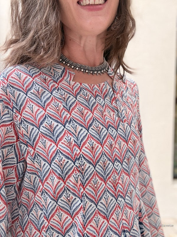 A close-up of a woman wearing a patterned long-sleeve top with a geometric leaf-like print in red, navy, and white, paired with a silver beaded choker necklace.