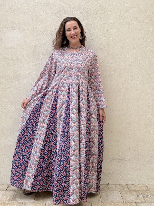 A woman modeling a long-sleeved maxi dress with a geometric pattern. The dress features a light pink and white diamond pattern on the upper bodice and sleeves, transitioning to a navy blue and pink floral pattern on the skirt, set against a textured beige stucco wall.
