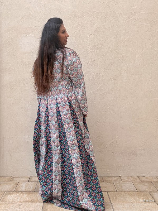 A woman wearing a long-sleeved maxi dress with a geometric pattern in pink, blue, and green, photographed from behind against a textured beige wall.