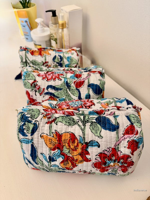 Three quilted cosmetic pouches with a colorful floral and botanical print in red, orange, blue, and green on a white background, arranged on a light-colored surface with toiletries in the background | Flower Bouquet