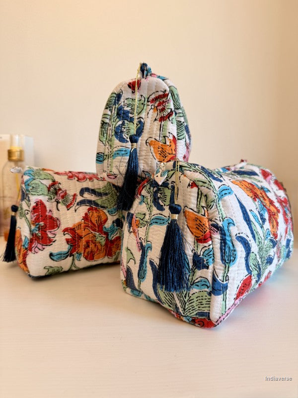 Three fabric cosmetic pouches with a colorful floral and botanical print in red, blue, green, and orange on a white background, featuring dark blue tassels, arranged on a light beige surface against a cream wall | Flower Bouquet
