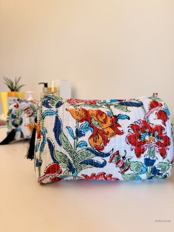 A cylindrical cosmetic pouch with a white background and a colorful floral print featuring red, orange, blue, and green flowers, shown on a beige surface with a blurred background | Flower Bouquet