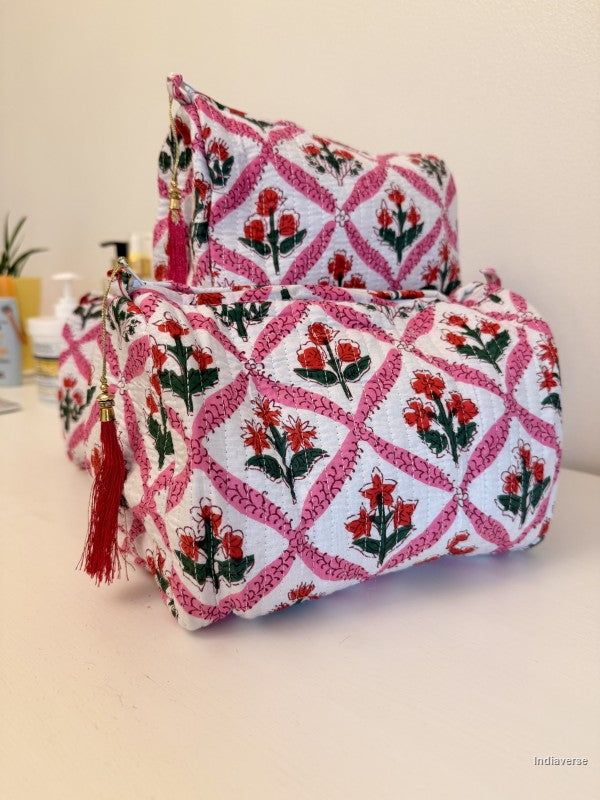 A set of two fabric cosmetic pouches stacked on a light beige surface against a cream wall. The pouches feature a white base with a pink diamond lattice pattern, red floral motifs, and red tassels | Flower Candy