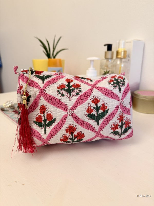 A small quilted clutch bag with a pink and white geometric lattice pattern, decorated with red and green floral motifs and a red tassel charm, resting on a light-colored tabletop with cosmetic bottles and a plant in the background | Flower Candy