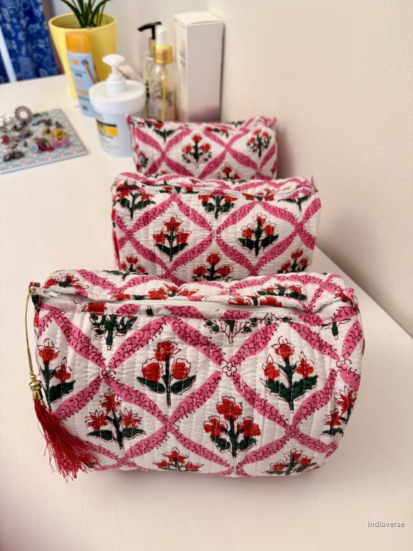 Three quilted fabric cosmetic pouches with a pink and red floral trellis pattern on a white background, arranged on a white tabletop with toiletries in the background | Flower Candy