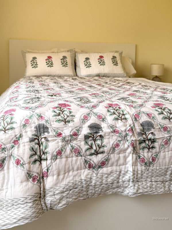 A quilted blanket with red and blue flower design arrange in a geometric pattern arranged on a white bed with two matching pillowcases