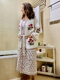 A woman wearing a long white embroidered robe with red floral motifs and a matching patterned skirt, standing in a tiled bathroom next to a bathtub and shelf.