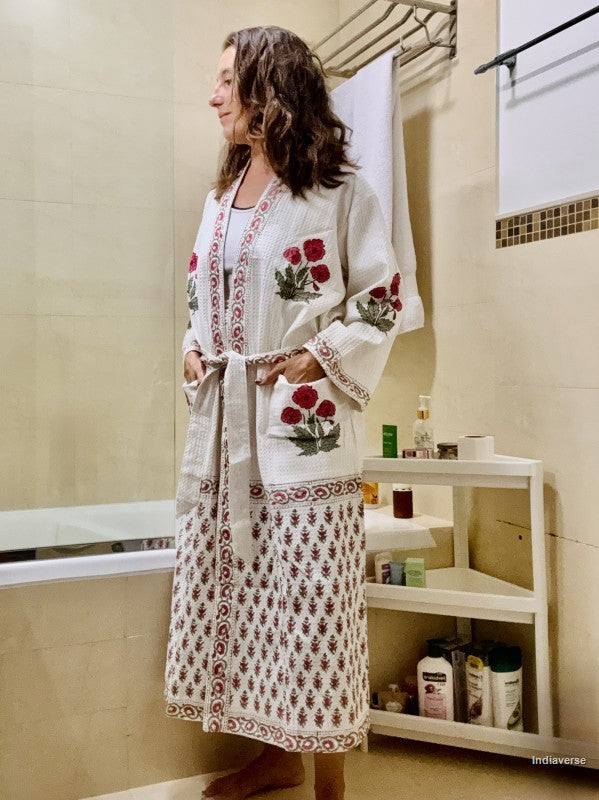 A woman wearing a long white embroidered robe with red floral motifs and a matching patterned skirt, standing in a tiled bathroom next to a bathtub and shelf.