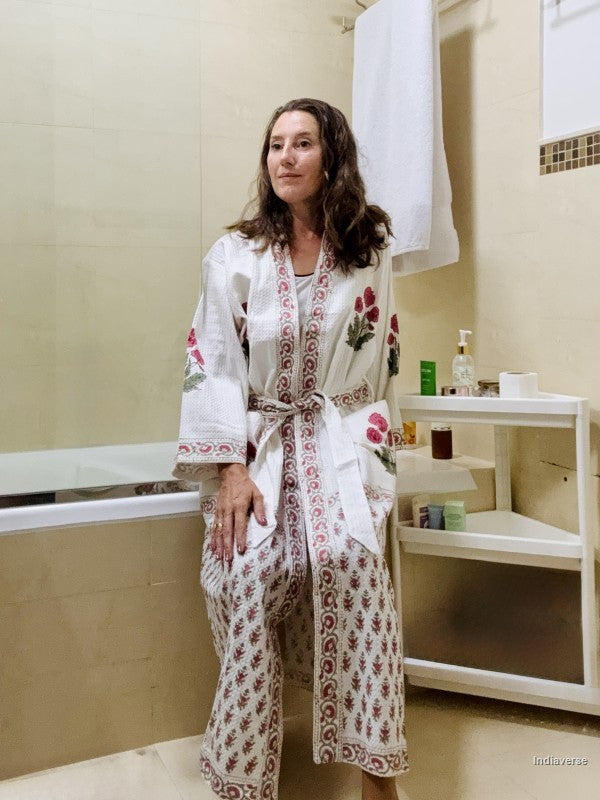 A woman wearing a white cotton robe with red and green floral embroidery and a matching patterned skirt, standing in a bathroom.
