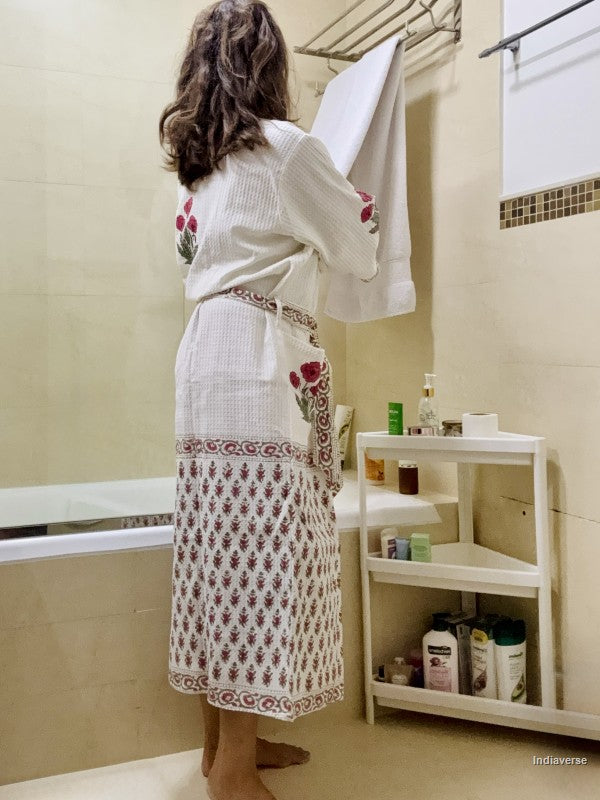 A woman stands with her back to the camera in a beige tiled bathroom, wearing a white cotton bathrobe with red floral embroidery and a matching patterned skirt. She is holding a white towel next to a bathtub and a white plastic storage shelf filled with toiletries.