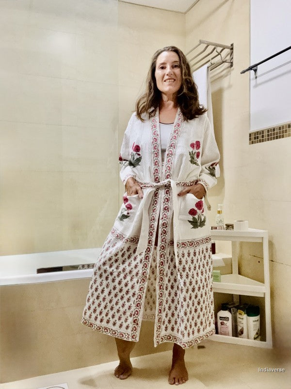 A woman wearing a white cotton bathrobe with red floral embroidery and a red geometric border pattern, standing in a beige tiled bathroom.