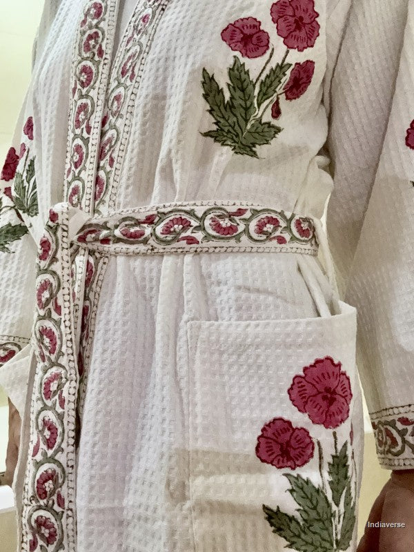 A close-up of a white textured cotton robe with pink floral embroidery and green leaves, featuring a matching embroidered waist belt and a front pocket.