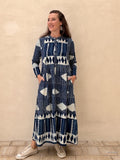 A woman modeling a long-sleeve maxi dress with a navy blue and white geometric tribal pattern, standing against a textured beige stucco wall.