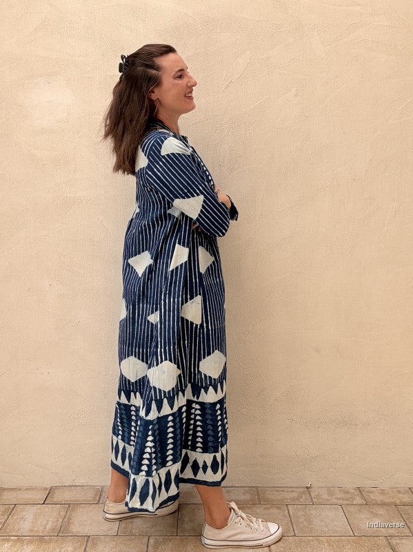 A woman wearing a long navy blue and white patterned maxi dress with vertical stripes and geometric motifs, standing against a textured beige wall.