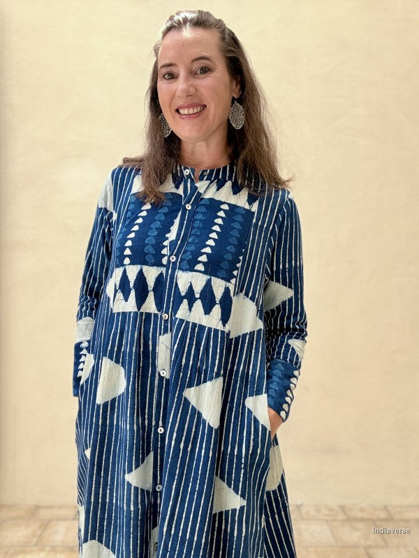 A woman wearing a long-sleeve button-front tunic dress with a blue and white geometric pattern, standing against a beige wall.