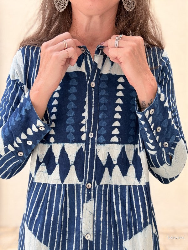 A close-up of a woman wearing a long-sleeve button-front top with a blue and white geometric block-print pattern. She is holding the collar open with both hands, wearing silver rings and earrings, against a neutral beige background.