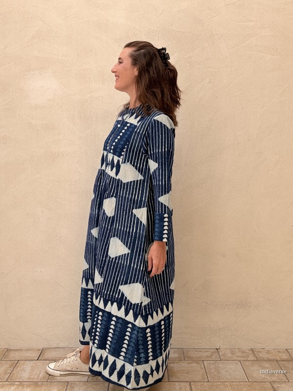 A woman wearing a long-sleeved navy blue and white patterned maxi dress, standing in profile against a textured beige wall.