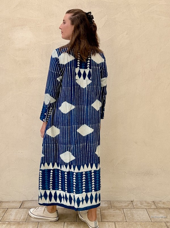 A woman wearing a long-sleeved navy blue and white patterned kaftan dress, shown from the back against a textured beige stucco wall.