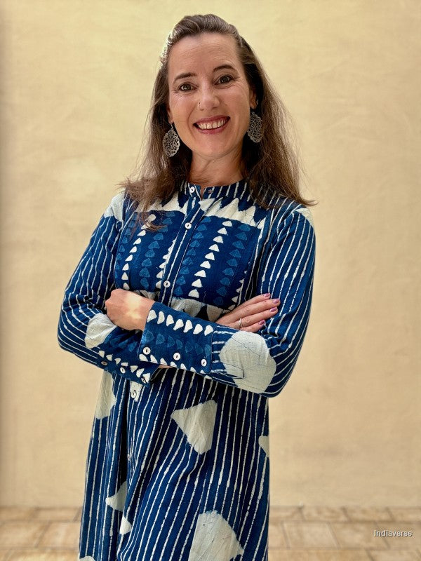 A woman with long brown hair is smiling and posing with her arms crossed, wearing a blue and white patterned long-sleeve tunic dress. She is standing against a textured beige wall.