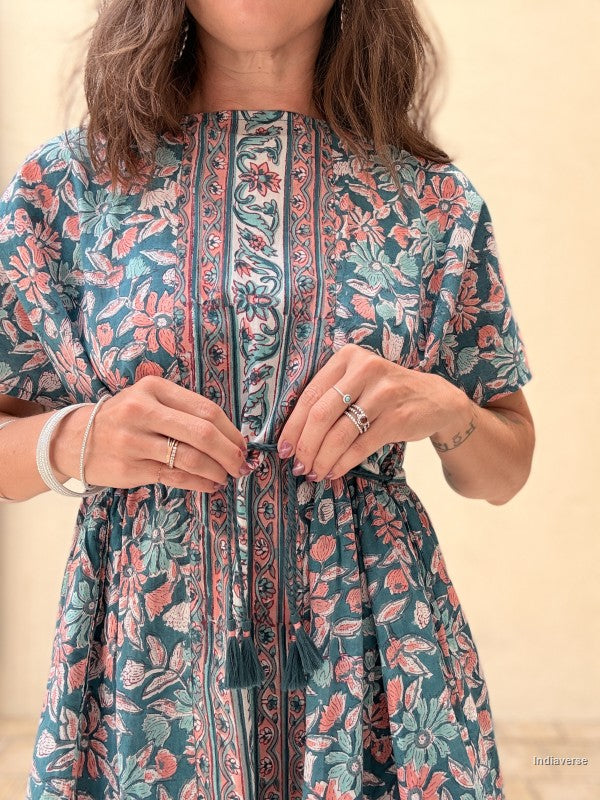 A close-up of a woman wearing a short-sleeve tunic dress with a teal blue-green base and a pink, coral, and sage green floral block print. She is adjusting the drawstring tie at the waist.