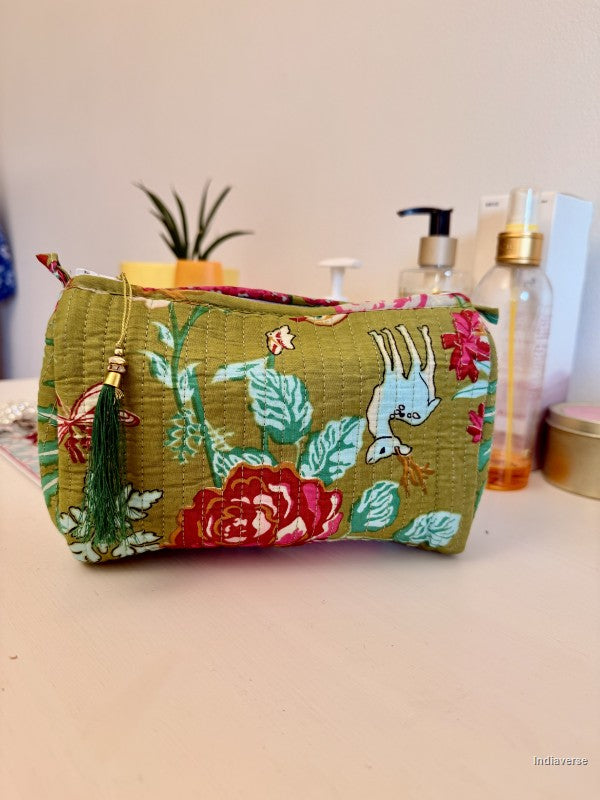 A fabric cosmetic pouch with a green base and a colorful floral print featuring red roses, green leaves, and a white bird. The pouch has a green tassel zipper pull and is shown sitting on a tabletop with toiletry bottles in the background | Magic Garden