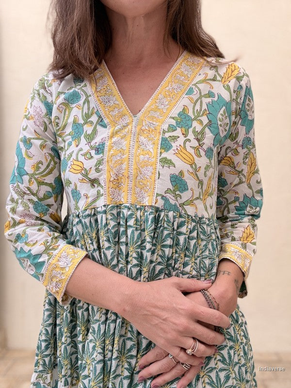 Close up of a woman wearing a long cotton dress with green palm tree motif and floral print standing against a beige wall