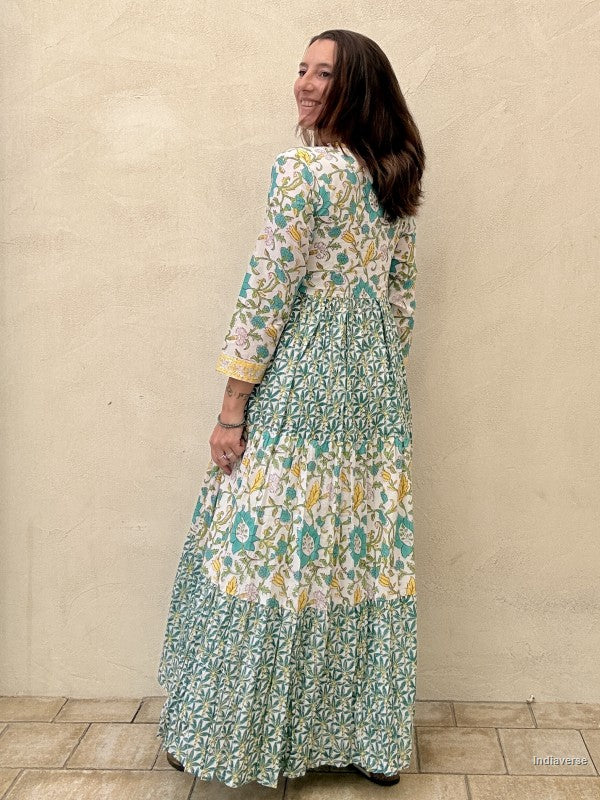 Backside view of a woman wearing a long cotton dress with green palm tree motif and floral print standing against a beige wall