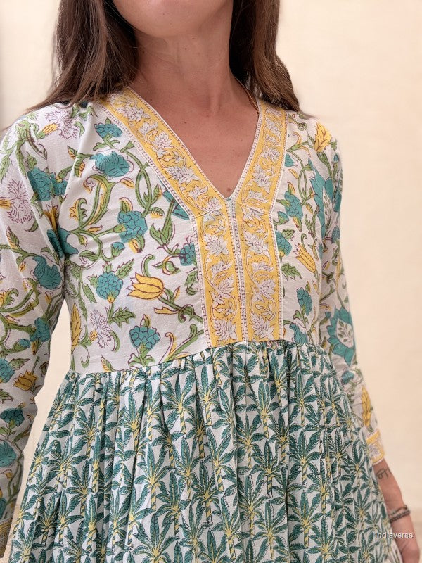 Close up of a woman wearing a long cotton dress with green palm tree motif and floral print standing against a beige wall