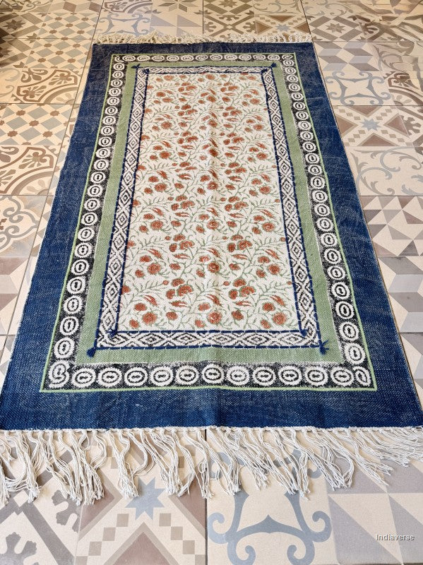 A rectangular Moroccan-style rug with a deep blue border, a light green inner border, and a cream center field decorated with a rust-orange floral pattern. The rug features white fringe along the bottom edge and is laid flat on a patterned tile floor.