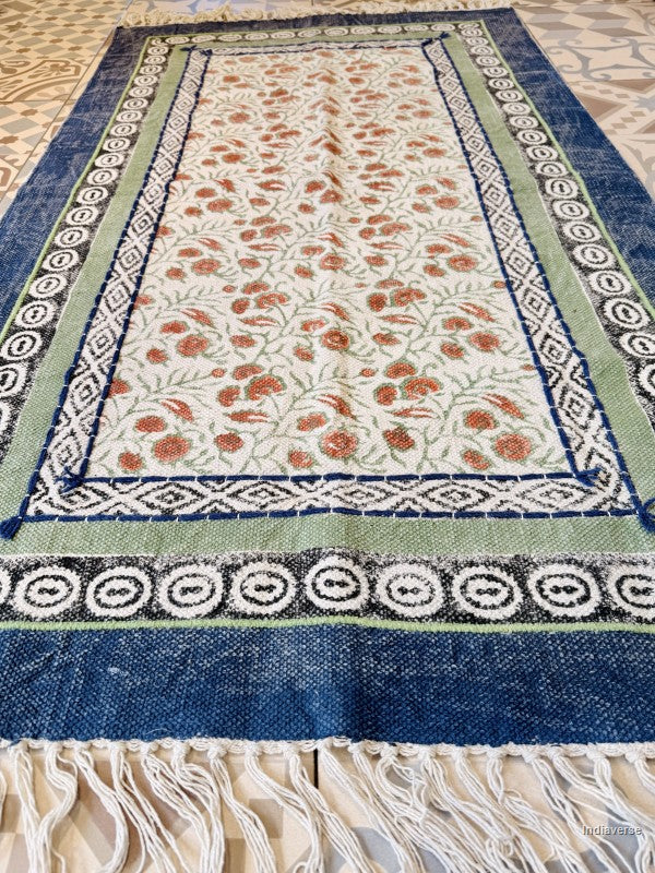 A rectangular woven rug with a cream center field decorated with orange and green floral motifs, surrounded by blue and green patterned borders, and finished with white fringe tassels.