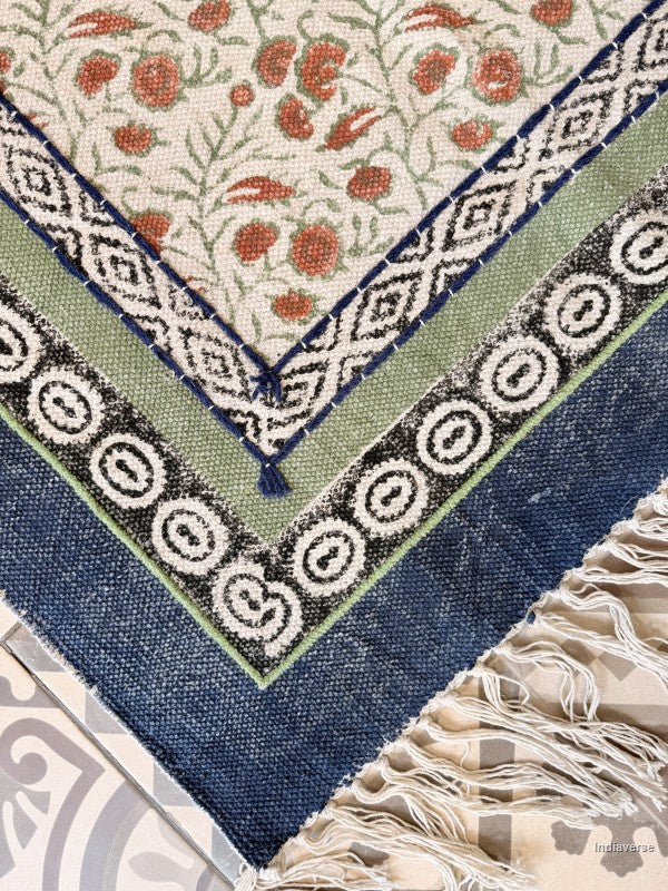 Close-up of a woven rug corner showing a cream field with rust-red floral motifs, layered border bands in navy, black, sage green, and cream, and off-white fringe.