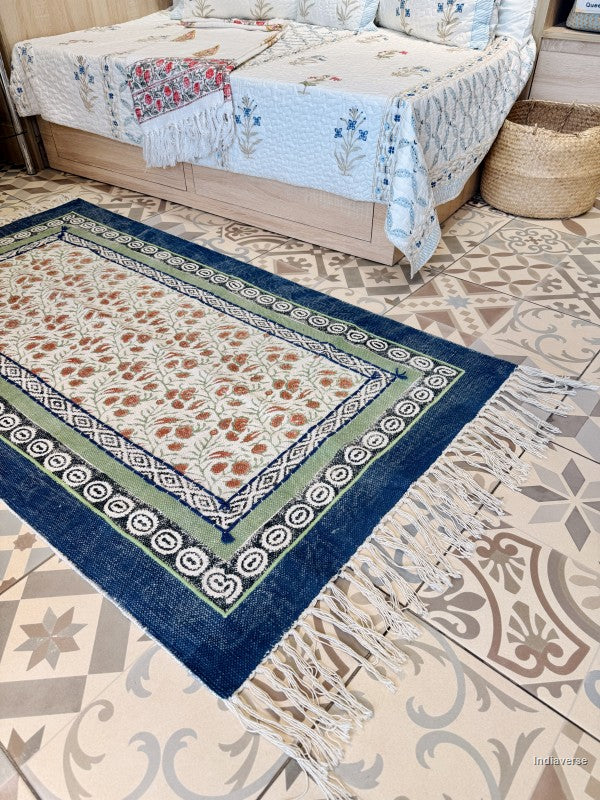 A rectangular area rug with a traditional bohemian pattern, featuring a navy blue border, green inner border, and cream center with floral motifs, shown on a patterned tile floor in a bedroom setting.