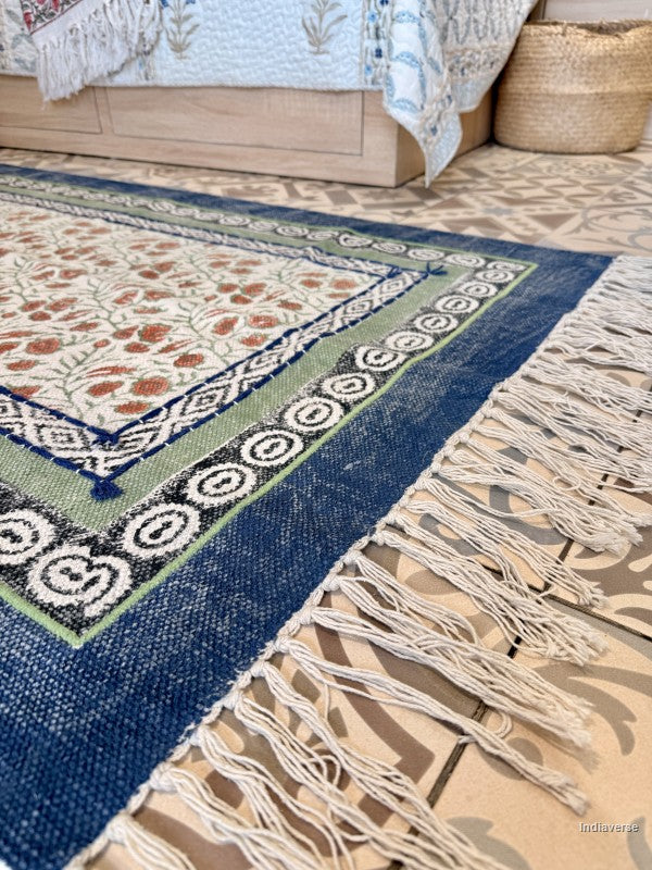 A close-up of a patterned area rug with a navy blue border, cream and beige central field, and white fringe tassels, shown on a patterned floor near a bed.