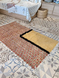 A rectangular area rug with a floral pattern in muted red, beige, and brown tones, featuring a solid mustard-yellow end panel bordered by black fringe, laid out on a patterned tile floor in a bedroom setting.